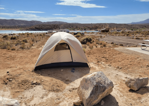 A tent set up on dry, rocky ground near a lake, with mountains and blue sky in the background.